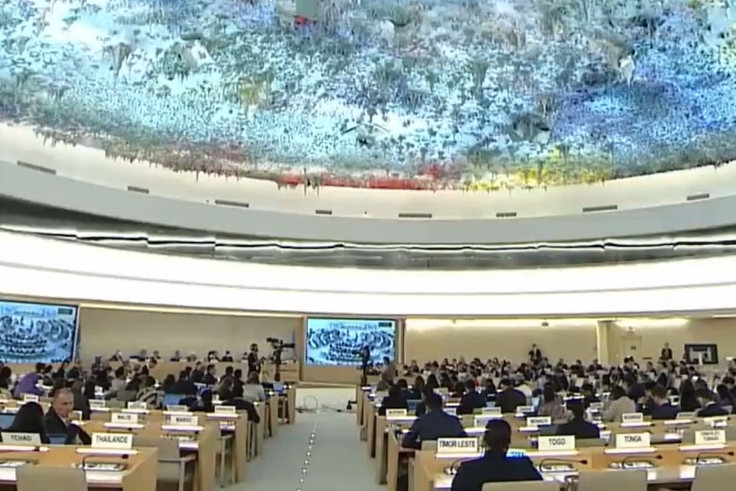 [ai] A large assembly hall with rows of delegates seated at wooden desks, some facing a screen displaying images. The ceiling features a colorful mural. The atmosphere appears formal, likely during an international conference or meeting.